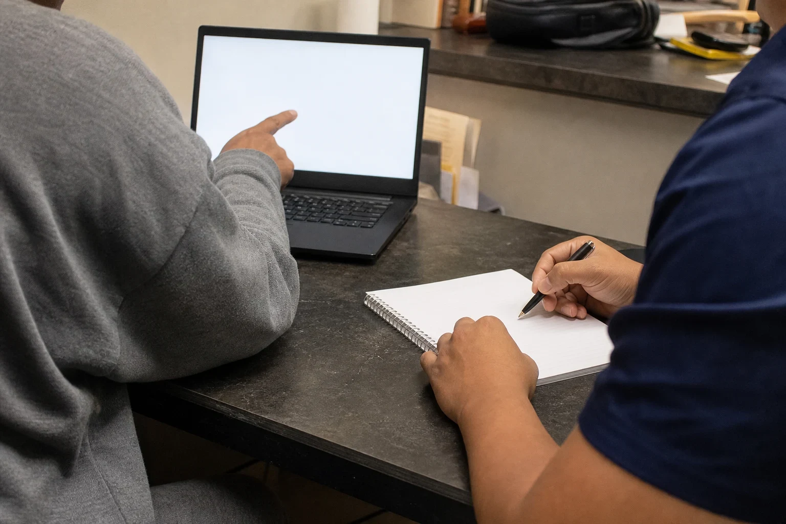 Two people at a dark office desk, one pointing at a laptop screen while the other writes notes in a spiral notebook