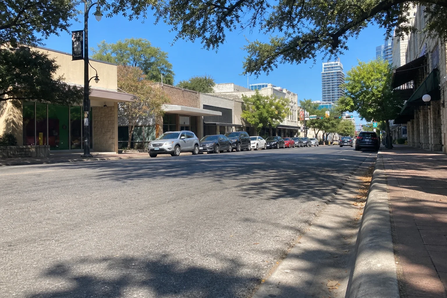 Low-angle view down a sunny downtown Austin street with parked cars along low storefronts, shade trees, a brick sidewalk and high-rise towers in the distance